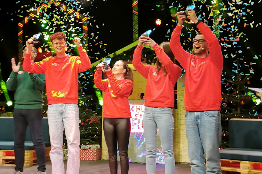 Vier Studierende stehen auf der Showbühne im Konfettiregen und halten ihre Pokale in die Luft.