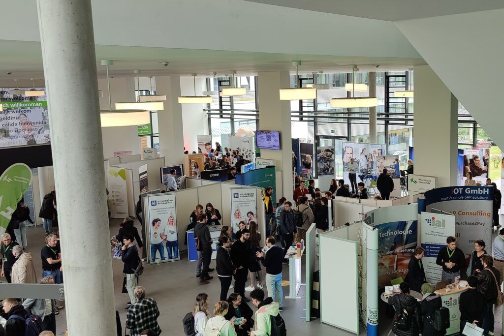 Im Foyer des Gelsenkirchener Campus sind Messestände diverser Unternehmen zu sehen, vor denen Studierende stehen.