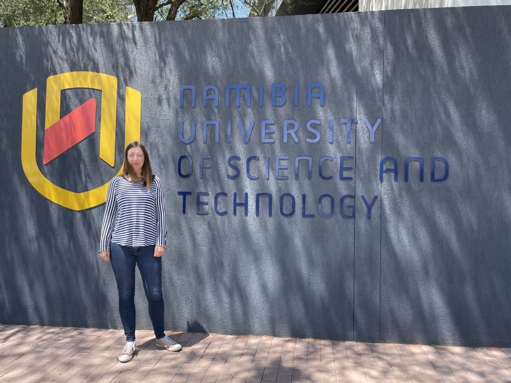 Katja Becker steht vor dem Eingang der Namibia University of Science and Technology vor einem großen Schriftzug der Universität.