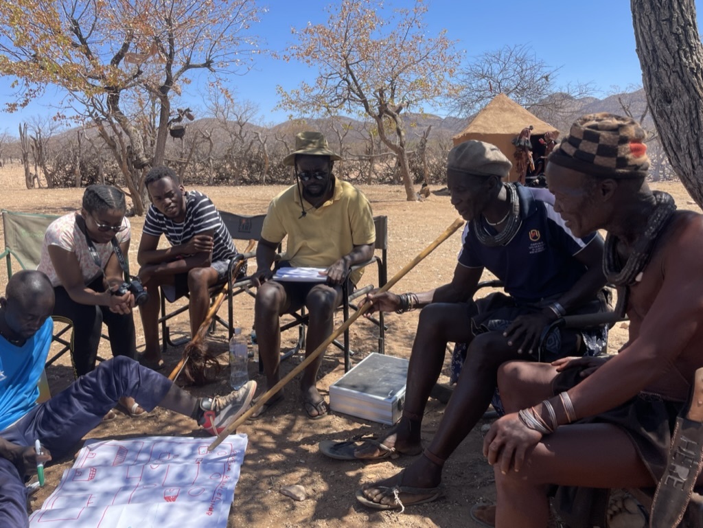 Universitätsbeschäftigte sitzen mit Menschen der Gruppe Ovahimba in ihrem Dorf bei einem Workshop.