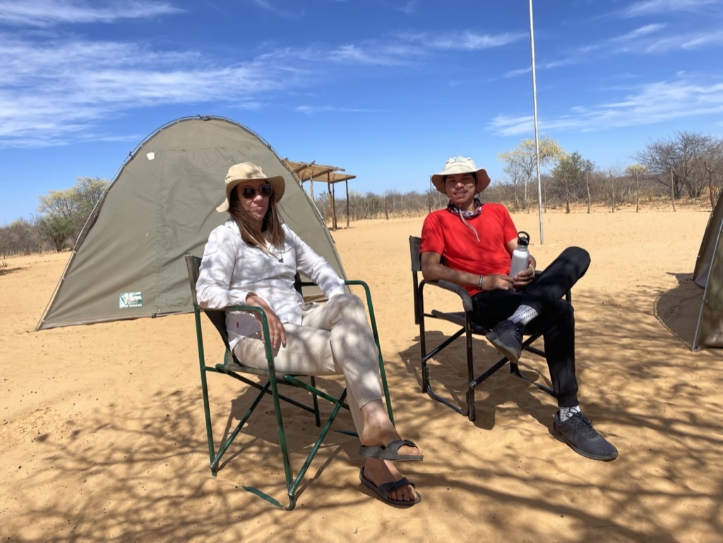 Katja Becker und Joseph-David Isaacs sitzen auf Campingstühlen vor einem aufgebauten Zelt im afrikanischen Busch.