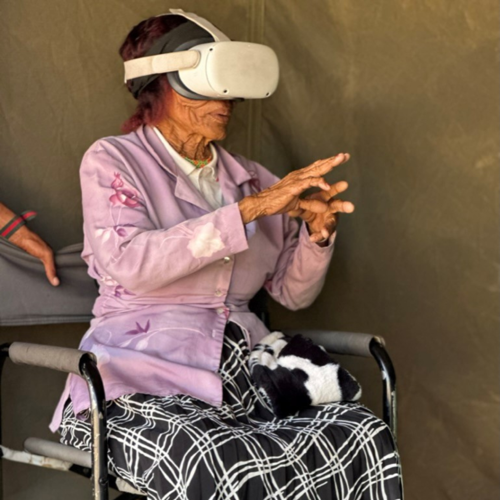 Eine ältere San-Frau sitzt auf einem Stuhl und trägt eine VR-Brille.