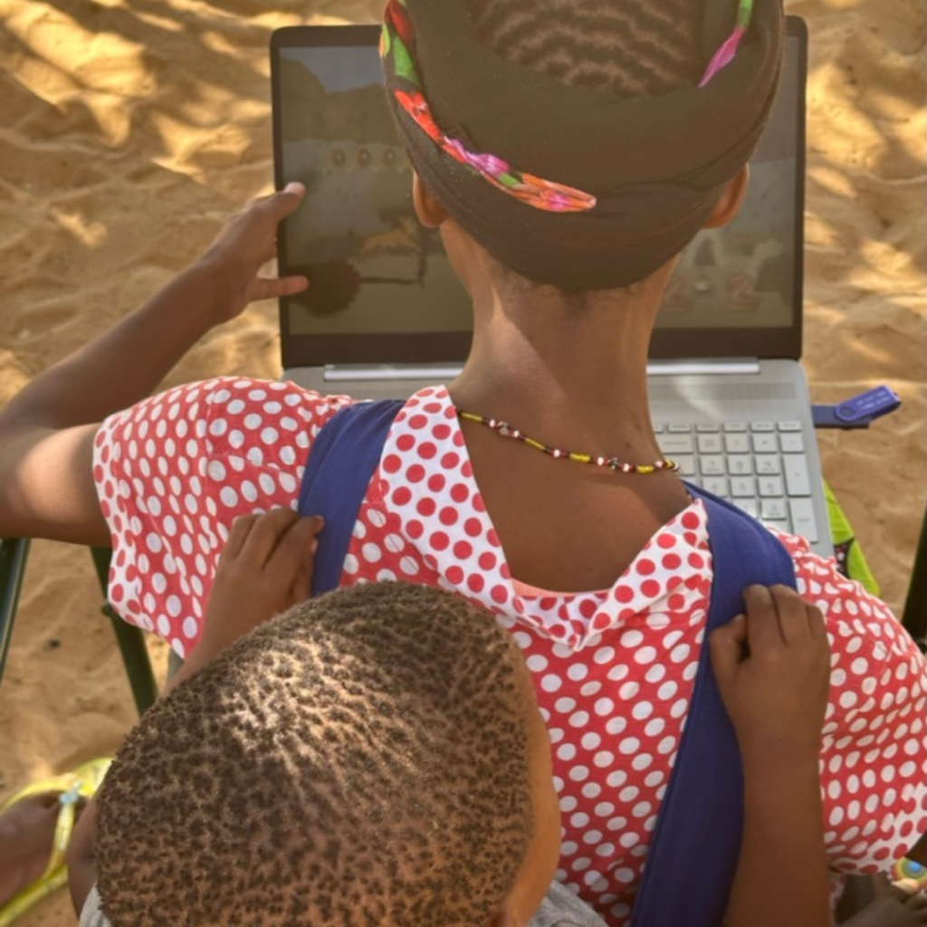 Eine junge San-Frau trägt ein Kleinkind auf ihrem Rücken und spielt ein VR-Spiel auf einem aufgeklappten Laptop.