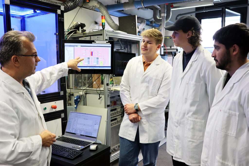 Drei Studierende stehen im Wasserstofflabor und Hören den Erklärungen eines wissenschaftlichen Mitarbeiters zu.