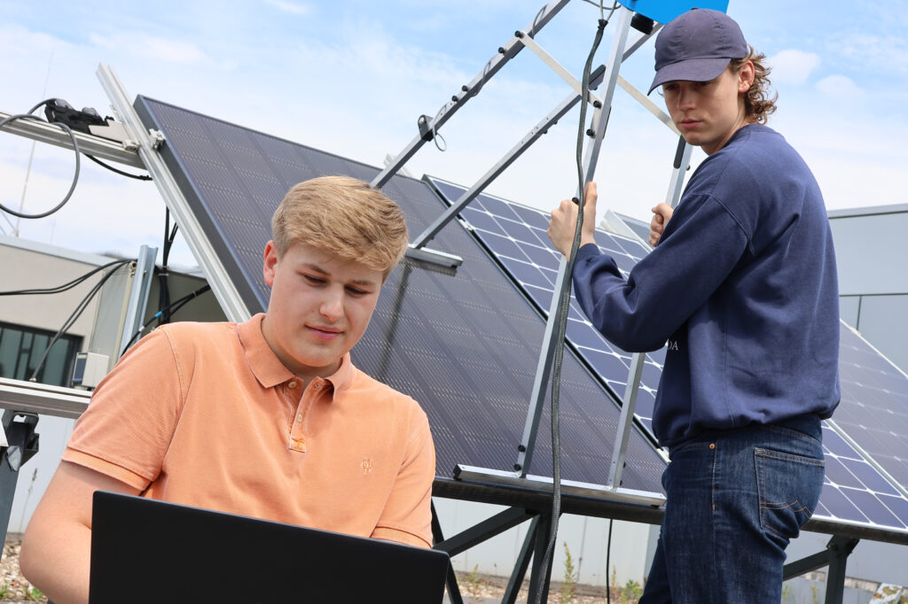 Zwei Studierende arbeiten an einem Solarpanel. Einer schaut auf sein Laptop, der andere steht vor dem Solarmodul.