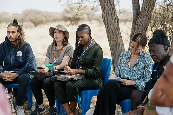 Teilnehmende des Workshops aus aller Welt sitzen im namibischen Busch auf Plastikstühlen.