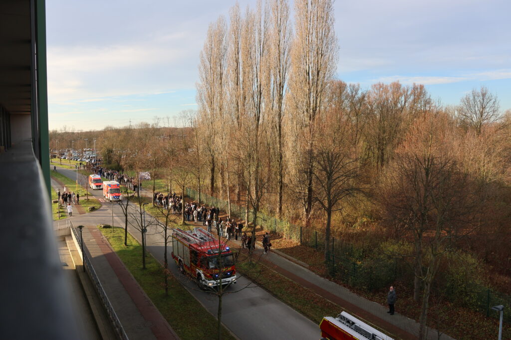 Blick aus dem Fenster auf die Straße. Im Hintergrund sieht man Mensche, die evakuiert werden. Gleichzeitig rollt die Feuerwehr an.