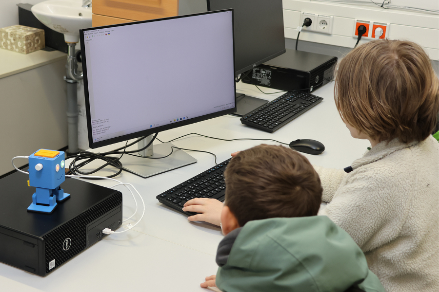 Zwei junge Schüler sitzen vor einem Computer, daneben steht ein kleiner blauer Roboter